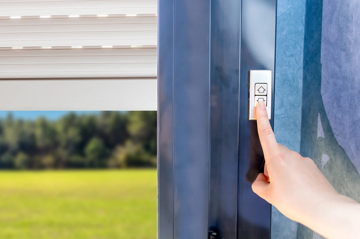 Une main actionne un bouton de commande électronique pour volet roulant, à proximité d'une baie vitrée donnant sur un jardin verdoyant